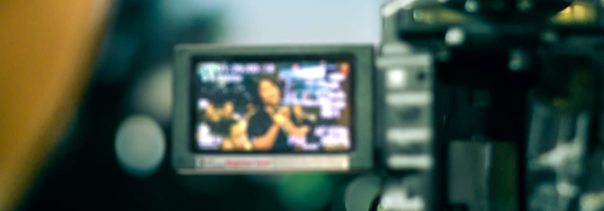 Nahaufnahme einer professionellen Videokamera, die eine Szene aufnimmt, in der eine Person spricht. Der LCD-Bildschirm der Kamera zeigt die Person scharf, während der Rest des Bildes unscharf ist, was den Videoanalyseprozess hervorhebt.