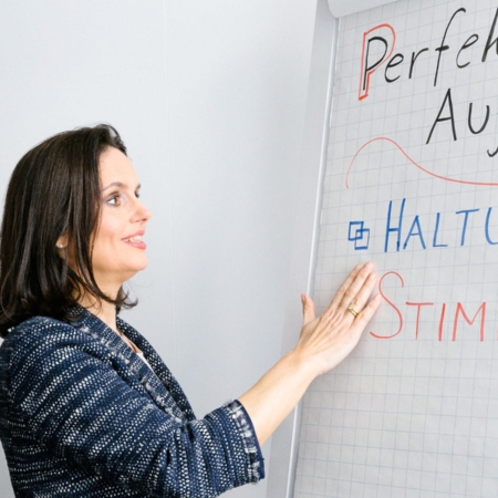 Eine Frau im blauen Blazer steht neben einem Flipchart und zeigt auf die handgeschriebenen Worte „Haltung“ und „Stimme“ an der Tafel. Das Flipchart scheint eine Präsentation über perfekte Leistung zu enthalten. Sie blickt auf die Tafel und lächelt leicht während des Seminars Workshop in Kleingruppe: Souveränes Auftreten und Präsentieren am 25.01.2023.