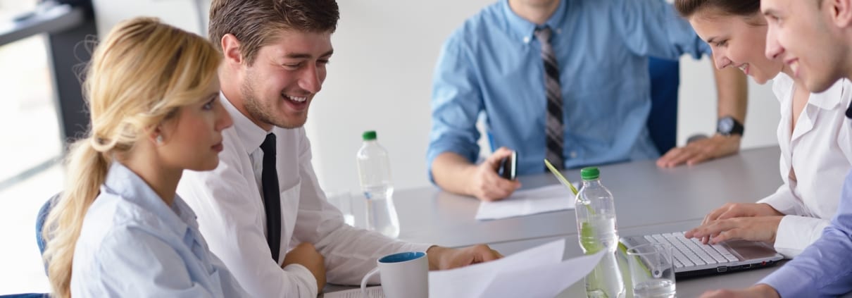 Eine Gruppe von fünf Personen sitzt in einem Büro um einen Tisch und diskutiert angeregt über „Wie gelingt Kommunikation“. Auf dem Tisch liegen Papiere, Notizbücher, ein Laptop und Wasserflaschen. Drei Männer und zwei Frauen sind anwesend, arbeiten zusammen und lachen gemeinsam.