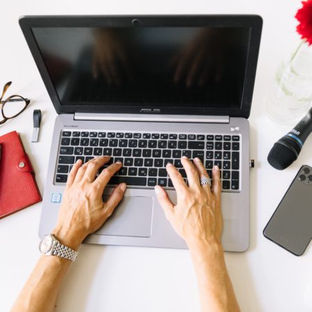 Eine Person tippt auf einem Laptop an einem weißen Schreibtisch. Daneben stehen ein rotes Notizbuch, eine Brille, ein Stift, ein iPhone und eine Tasse Kaffee. Eine leuchtend rote Blume in einer Vase bringt Farbe in den Arbeitsplatz. Auf dem Bildschirm steht „In wenigen Schritten zur gelungenen Pressemitteilung“, was darauf hindeutet, dass der Fokus auf dem Verfassen von Pressemitteilungen liegt.