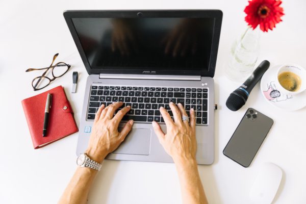 Eine Person tippt auf einem Laptop an einem weißen Schreibtisch. Daneben stehen ein rotes Notizbuch, eine Brille, ein Stift, ein iPhone und eine Tasse Kaffee. Eine leuchtend rote Blume in einer Vase bringt Farbe in den Arbeitsplatz. Auf dem Bildschirm steht „In wenigen Schritten zur gelungenen Pressemitteilung“, was darauf hindeutet, dass der Fokus auf dem Verfassen von Pressemitteilungen liegt.