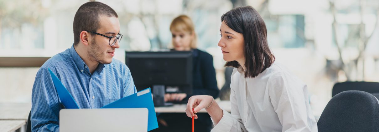 Zwei Kollegen sitzen an einem Schreibtisch in einem Büro und diskutieren 10 Tipps für gute Kommunikation. Einer hält einen blauen Ordner und hat einen Laptop vor sich, während der andere ein Blatt Papier und einen roten Stift hält. Eine weitere Person arbeitet im Hintergrund an einem Computer und trägt zum allgemeinen Kommunikationserfolg bei.