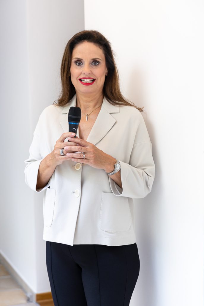 Eine Frau mit langen braunen Haaren, in einem hellen Blazer und einer schwarzen Hose steht lächelnd mit einem Mikrofon da – bereit für den „Power-Workshop: Fit und sicher für den nächsten Auftritt am 04.09.2025“.