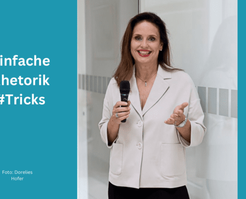 Eine Frau steht da und lächelt in die Kamera, während sie ein Mikrofon hält und eine Handbewegung macht, um ihre Überzeugungskraft zu demonstrieren. Der Text links lautet „Einfache Rhetorik #Tricks“ mit einem Fotonachweis an „Doreilles Hofer“ darunter. Der Hintergrund ist ein heller Innenraum.