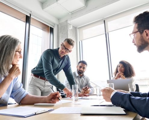 Eine Gruppe von fünf Personen nimmt an einem Team-Workshop in einem modernen Büro teil. Eine Person steht und deutet auf Dokumente auf dem Tisch, während die anderen sitzen und aufmerksam zuhören. Ein Laptop und Papiere sind auf dem Tisch ausgebreitet, im Hintergrund sind große Fenster zu sehen.