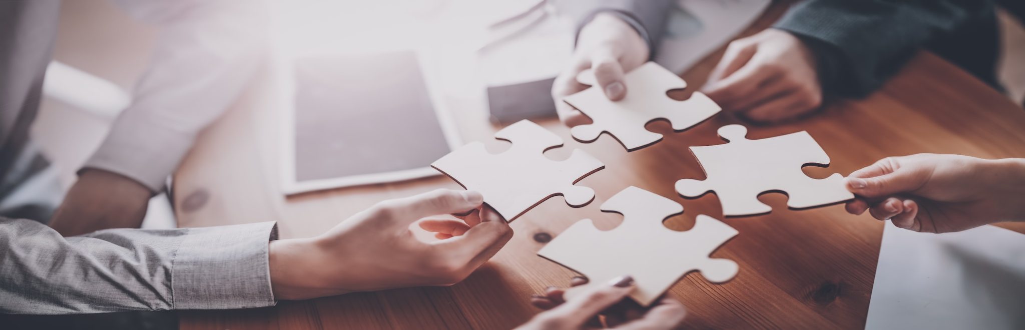 Vier Hände, die große weiße Puzzleteile über einem Holztisch verbinden, symbolisieren die Zusammenarbeit in Teamworkshops. Im Hintergrund liegt ein Tablet auf dem Tisch und verstärkt den Eindruck gemeinsamer Anstrengung.