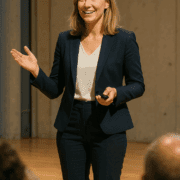 Eine Frau in einem marineblauen Anzug und weißer Bluse steht in einem Raum, lächelt und gestikuliert mit einer Hand, während sie eine Fernbedienung hält, und scheint eine Präsentation mit einer Checkliste zur Vorbereitung einer Präsentation zu halten.