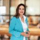 Eine Frau mit langen braunen Haaren, die einen türkisfarbenen Blazer und eine weiße Bluse trägt, steht mit gefalteten Händen in einem Haus und lächelt in die Kamera. Der Hintergrund ist sanft verschwommen und warm beleuchtet.