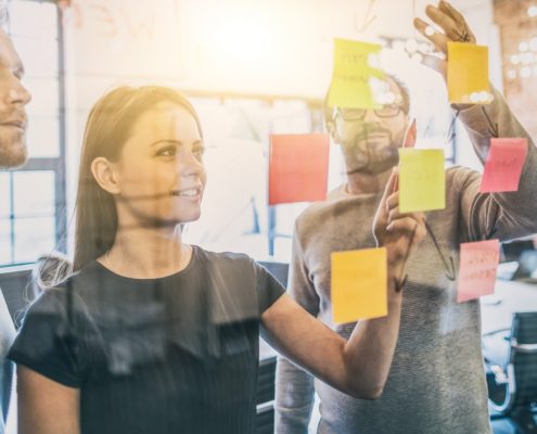 Drei Personen arbeiten in einem modernen Büro an einer Glaswand, die mit bunten Haftnotizen bedeckt ist. Sie diskutieren und brainstormen Ideen bei natürlichem Sonnenlicht während eines Workshops: Moderieren mit Leichtigkeit.