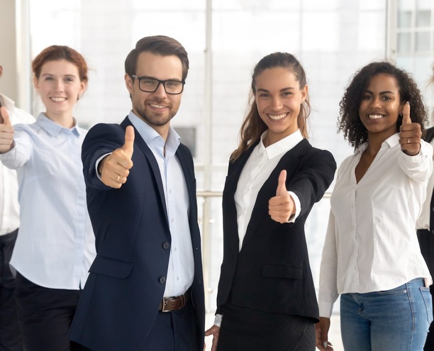 Eine Gruppe von sechs verschiedenen Fachleuten, drei Männer und drei Frauen, stehen in einem Innenraum zusammen, lächeln und zeigen Daumen nach oben. Sie zeigen empathische Führungskommunikation in einer hellen Büroumgebung und tragen Geschäftskleidung.