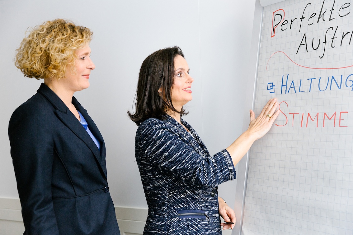 Zwei Frauen in Business-Kleidung stehen während eines Seminars Abenteuer Kommunikation an einem Flipchart. Eine zeigt auf einen Text, auf dem "Perfekte Auftritte", "Haltung" und "Stimme" steht. Beide lächeln und sind in eine lebhafte Diskussion vertieft.
