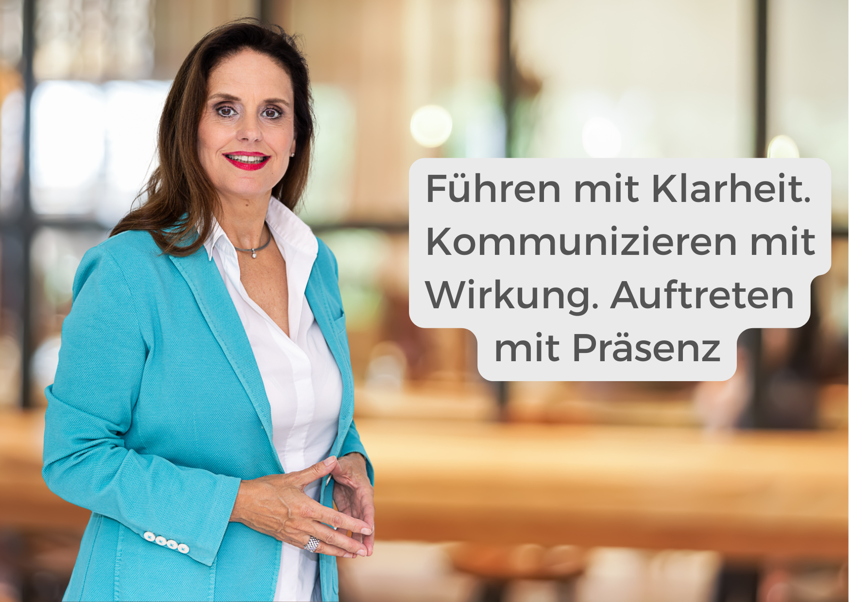 Eine Frau in einem türkisfarbenen Blazer steht in einem Haus und lächelt in die Kamera. Neben ihr steht ein deutscher Text: "Führen mit Klarheit. Kommunizieren mit Wirkung. Auftreten mit Präsenz."-Teil des Seminars Abenteuer Kommunikation.